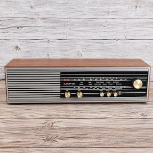 Può includere: Una radio rettangolare Europhon con finitura effetto legno, dotata di un pannello frontale nero con strisce argentate orizzontali, un display delle frequenze con nomi di città e cinque manopole di controllo circolari dorate.