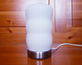 May include: A table lamp featuring a cylindrical, white frosted glass shade with a distinct three-tiered, rounded, bulbous silhouette. The lamp is supported by a circular, brushed silver-tone metal base and includes a white power cord.