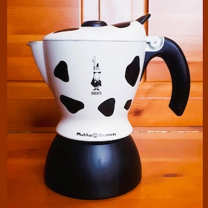 Könnte beinhalten: Eine weiße und schwarze Espressomaschine mit Kuhmuster, schwarzem Griff und schwarzem Sockel. Die Maschine trägt den Schriftzug "Bialetti" und "Mukka Express".