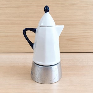 Pode incluir: Uma cafeteira italiana de cerâmica branca e azul. A alça e o botão da tampa são azuis escuros. A base é de metal prateado. A cafeteira tem um design clássico.