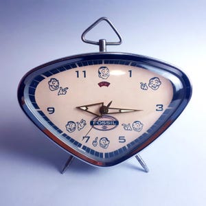 May include: A retro Fossil alarm clock with a cream face and a distinctive triangular design. The clock has a silver-tone metal frame, a handle, and three legs. The face features cartoon illustrations and the Fossil logo.