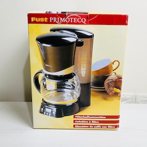 Op de afbeelding: Een zwarte en gouden koffiezetapparaat met een glazen kan en een digitaal display. De doos heeft de merknaam "Fust Primotec" en de tekst "Filterkaffeemaschine, Cafetière à filtre, Macchina da caffe con filtro".