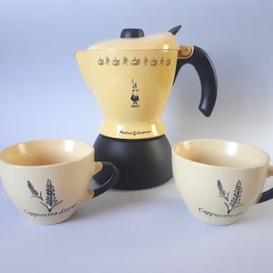 Könnte beinhalten: Eine cremefarbene Bialetti Mukka Express Herdkanne mit schwarzem Boden und Griff, verziert mit einem Muster aus kleinen Kaffeetassen und Logo. Zwei passende cremefarbene Keramiktassen mit schwarzen Henkeln und einem Weizenmotiv Cappuccino d'orzo.