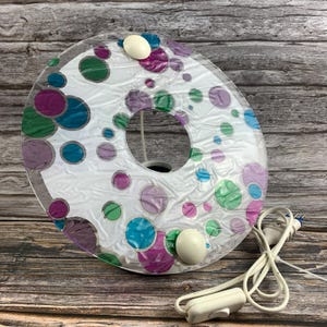 May include: A circular wall lamp featuring a white background decorated with scattered purple, teal, and green circles of varying sizes. The lamp has a translucent, crinkled plastic cover, two white mounting knobs, and a white power cord with a switch.