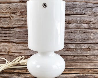 May include: A white glass table lamp featuring a cylindrical shade and a rounded, bulbous base. A small circular sticker with black text is visible on the upper portion of the shade. A white electrical cord with a switch is attached to the base.