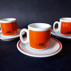 May include: Three espresso cups and saucers. The cups are orange with white interiors and handles. Each cup has the word "HAG" with a heart symbol. The saucers are white with an orange rim.