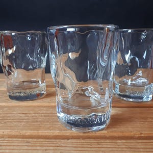 May include: Three clear, textured glass tumblers with a wavy design. The glasses are cylindrical with a slightly irregular rim and base. The glasses are on a wooden surface.