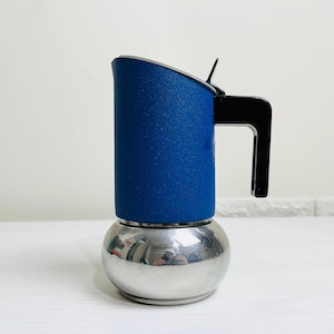 Pode incluir: Uma cafeteira italiana com um corpo superior cilíndrico azul de acabamento texturizado e uma base arredondada de aço inoxidável polido. O dispositivo inclui uma pega de plástico preto e uma tampa articulada com uma pequena alavanca metálica.