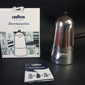 Pode incluir: Uma cafeteira espresso Lavazza Carmencita Classic em aço inoxidável com uma alça preta e uma caixa com o mesmo design. A caixa tem o logotipo da Lavazza e as palavras "Carmencita Classic" impressas.
