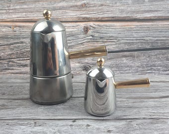 May include: Two stainless steel stovetop espresso makers with gold-toned handles and knobs. The larger coffee maker is approximately 8 inches tall, and the smaller one is about 4 inches tall. Both are on a wooden surface.