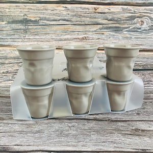 Może przedstawiać: Zestaw sześciu jasnoszarych ceramicznych filiżanek do espresso o charakterystycznej, pogniecionej fakturze. Filiżanki są ułożone w dwóch rzędach po trzy sztuki i umieszczone w przezroczystej plastikowej wytłoczce z widocznym czarnym napisem.