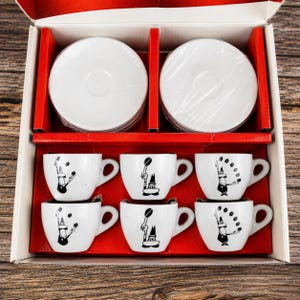 Pode incluir: Um conjunto de seis chávenas de café em cerâmica branca com pires a condizer numa caixa de cartão. Cada chávena apresenta um gráfico preto de uma figura estilizada a fazer malabarismos com grãos de café. Os pires estão envolvidos em plástico.
