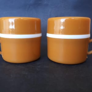 Op de afbeelding: Twee oranje mokken met een witte band in het midden. Elke mok heeft een klein, gebogen oor. De mokken zijn gemaakt van een glanzend materiaal en staan op een donkere ondergrond. De mokken zijn een bijpassende set.
