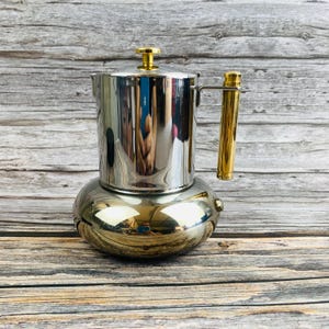 Puede incluir: Una cafetera de acero inoxidable pulido con una base ancha y redondeada y una cámara superior cilíndrica. Cuenta con un asa de metal dorado y un pomo a juego en la tapa, lo que proporciona un acabado metálico y altamente reflectante.