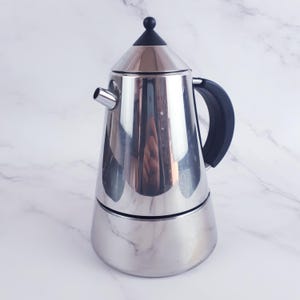 Op de afbeelding: Een roestvrijstalen espressomaker met een zwarte handgreep en een zwarte knop bovenop. De koffiezetter heeft een gepolijst, reflecterend oppervlak en een tuit om uit te schenken. Het ontwerp is een klassieke Italiaanse stijl.