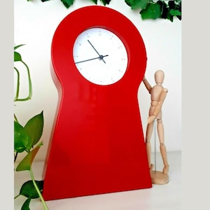 May include: A striking red clock with a white face and black hands, featuring a distinctive, rounded shape. A wooden artist's mannequin stands next to the clock. Green foliage is visible in the background.