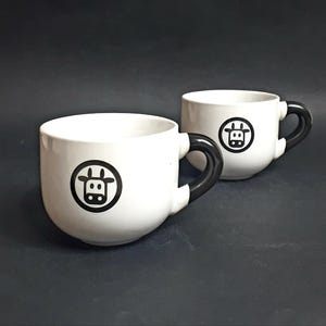 Puede incluir: Dos tazas de cerámica blanca con asas negras y un diseño de silueta de vaca negra.