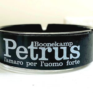 Peut inclure: Cendrier noir avec un bord argenté. Il porte les mots "Petrus l'amaro per l'uomo forte" et "Boonekamp" imprimés en blanc.