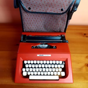 Peut inclure: Une machine à écrire Olivetti College rouge avec des touches blanches et un chariot noir. La machine à écrire est ouverte, révélant les touches et l'alimentation du papier. L'étui de transport est marron avec un motif.