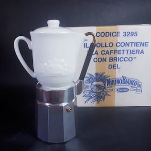 Puede incluir: Una taza de café de cerámica blanca con tapa se encuentra sobre una cafetera espresso plateada y negra. El asa es negra con un acento dorado. Una caja con texto italiano está en el fondo.
