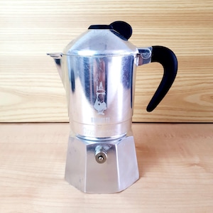 Op de afbeelding: Een zilveren espressomaker voor op het fornuis met een zwarte handgreep en bovenkant. De koffiemaker heeft een klassiek ontwerp met een kenmerkende vorm en een klein logo op de voorkant. Hij staat op een houten oppervlak.