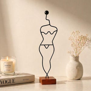 May include: A black metal sculpture of a female form, mounted on a small wooden base. The sculpture has a minimalist design, with a rounded head and a flowing, abstract body shape. A candle in a glass jar and a book are in the background.