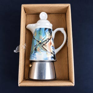 Pode incluir: Uma cafeteira italiana com uma parte superior de cerâmica branca com uma estampa gráfica abstrata colorida, uma tampa branca com puxador e uma base de metal prateado. A cafeteira está colocada dentro de uma caixa de papelão ondulado.
