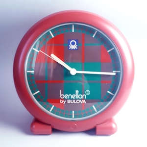 Könnte beinhalten: Roter Benetton by Bulova Wecker. Das Zifferblatt zeigt ein rot-grünes Karomuster mit weißen Zeigern und Markierungen. Das Benetton-Logo ist sichtbar. Klassisches Design.