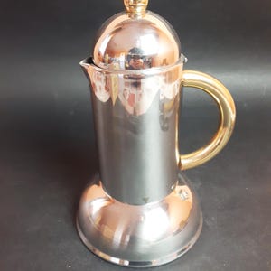 Peut inclure: Une cafetière en argent et or avec une base arrondie et un couvercle avec un bouton sur le dessus. La poignée est dorée.
