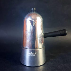 Pode incluir: Uma cafeteira italiana prateada com uma alça preta e um botão preto na parte superior. A marca "Lavazza" está impressa na base. A cafeteira é feita de metal e foi projetada para preparar café.