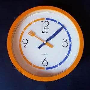 Op de afbeelding: Oranje en witte quartz klok met een vorkvormige uurwijzer. De klok heeft blauwe en oranje accenten, met het woord "bino" erboven. De diameter van de klok is ongeveer 20 cm.