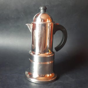 May include: A stainless steel stovetop espresso maker with a black handle and a black knob on top. The coffee maker has a classic design and is made of shiny metal. The handle is curved for easy gripping.