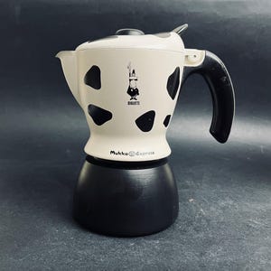 Puede incluir: Una cafetera de espresso Bialetti Mukka Express blanca y negra con una ilustración de dibujos animados de un hombre con sombrero. La cafetera tiene un asa negra y una base negra.