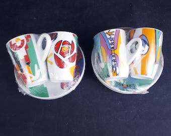 May include: Two sets of white ceramic cups and saucers wrapped in clear plastic. The left set features red, green, and yellow geometric patterns. The right set displays a colourful design with yellow, blue, and purple abstract shapes and figures.