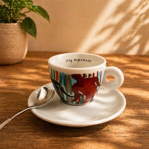 Op de afbeelding: Een witte keramische espressokop en schotel met een kleurrijk abstract ontwerp van rode, groene en blauwe verticale strepen en een donkerrode figuur. De binnenrand van de kop is bedrukt met de tekst illy espresso. Een zilveren lepel in hartvorm.