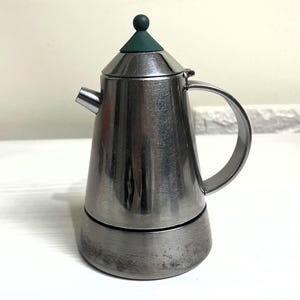 Pode incluir: Uma cafeteira de espresso de metal prateado com uma tampa de plástico verde. A cafeteira tem um corpo arredondado e uma alça.