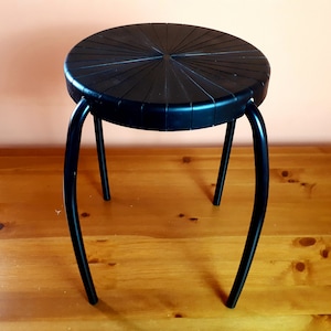Könnte beinhalten: Ein schwarzer dreibeiniger Hocker mit einer runden Sitzfläche. Die Sitzfläche hat ein Sonnenstrahlen-Design.