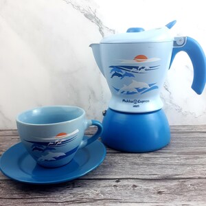 Könnte beinhalten: Ein hellblaues Espresso-Set mit Kaffeemaschine, Tasse und Untertasse. Die Espressomaschine und die Tasse zeigen ein Design von Delfinen, die im Meer springen, mit einer untergehenden Sonne. Auf der Espressomaschine steht "Mukka Express".