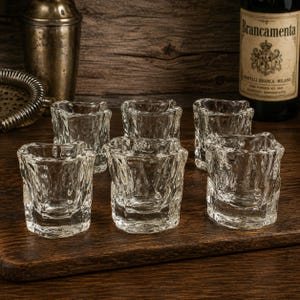 Può includere: Un set di sei bicchierini da shot in vetro trasparente con una superficie strutturata, irregolare e organica. Ogni bicchiere ha una base spessa e un bordo leggermente irregolare, creando un aspetto grezzo. I bicchieri sono disposti in gruppo su una superficie scura.