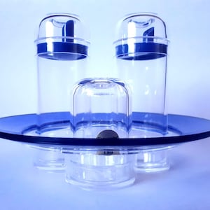 Puede incluir: Un juego de servir de vidrio transparente con detalles en azul. El conjunto incluye dos recipientes cilíndricos altos con tapas azules, un pequeño recipiente en forma de cúpula y una bandeja redonda. Diseñado para servir alimentos o bebidas.