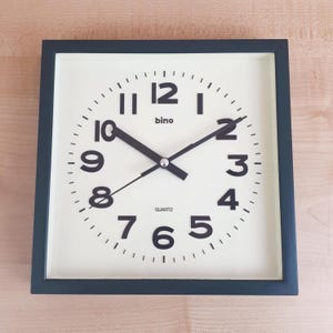 Könnte beinhalten: Quadratische Wanduhr mit schwarzem Rahmen und cremefarbenem Zifferblatt. Die Uhr hat schwarze Ziffern, Zeiger und das Wort "bino" oben. Das Wort "QUARTZ" ist unten aufgedruckt. Klassisches Design.