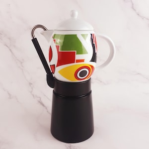Pode incluir: Uma cafeteira italiana preta com um topo de cerâmica colorido. O topo de cerâmica tem um fundo branco com formas geométricas vermelhas, verdes e amarelas. A alça é prateada e preta.