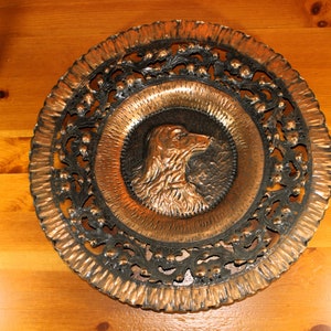 Op de afbeelding: Een koperen wandplaat met een decoratief, zwart, uitgesneden ontwerp rond de rand. Het midden van de plaat heeft een reliëf, koperen afbeelding van een hondenkop.