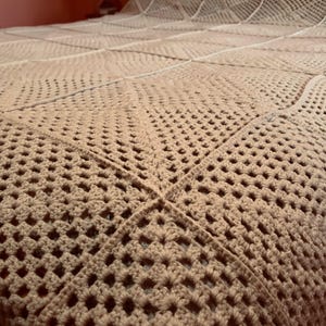 Könnte beinhalten: Eine braune Häkeldecke mit quadratischem Muster. Die Decke ist aus dickem Garn gefertigt und hat eine weiche, kuschelige Textur.