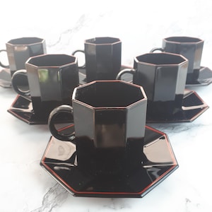 Peut inclure: Un ensemble de six tasses à thé et soucoupes en céramique noire. Chaque tasse a une forme octogonale avec une anse. Les soucoupes sont carrées avec une bordure rouge. L'ensemble est présenté sur une surface blanche.