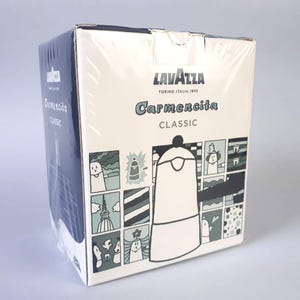 May include: A rectangular cardboard box for a Lavazza Carmencita Classic coffee maker. The white packaging features dark blue text and a grid of illustrated panels in shades of blue, green, and white, depicting coffee-related scenes and motifs.