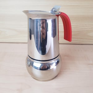 Pode incluir: Uma cafeteira de aço inoxidável polido com base arredondada e câmara superior cilíndrica. A cafeteira possui uma alça vermelha brilhante e um mecanismo de dobradiça de metal prateado na tampa, com uma pequena válvula na lateral.
