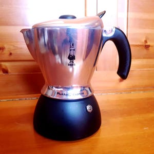 Könnte beinhalten: Ein silberner und schwarzer Espressokocher für den Herd mit einem schwarzen Griff. Der Kaffeezubereiter hat ein klassisches Design mit dem Markennamen "Bialetti" und "Mokka Express". Er steht auf einer Holzoberfläche.