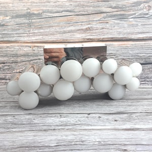 Può includere: Un moderno apparecchio di illuminazione per vanità con finitura cromata e più lampadine bianche a forma di globo. Le lampadine sono disposte a grappolo, creando una luce soffusa e diffusa. L'apparecchio è progettato per l'uso in bagno o in camera da letto.