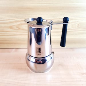 Può includere: Una caffettiera in acciaio inossidabile con manico e pomello neri. La caffettiera ha una base rotonda e una camera superiore cilindrica. Il logo del marchio è visibile sulla parte anteriore. Progettata per preparare il caffè.